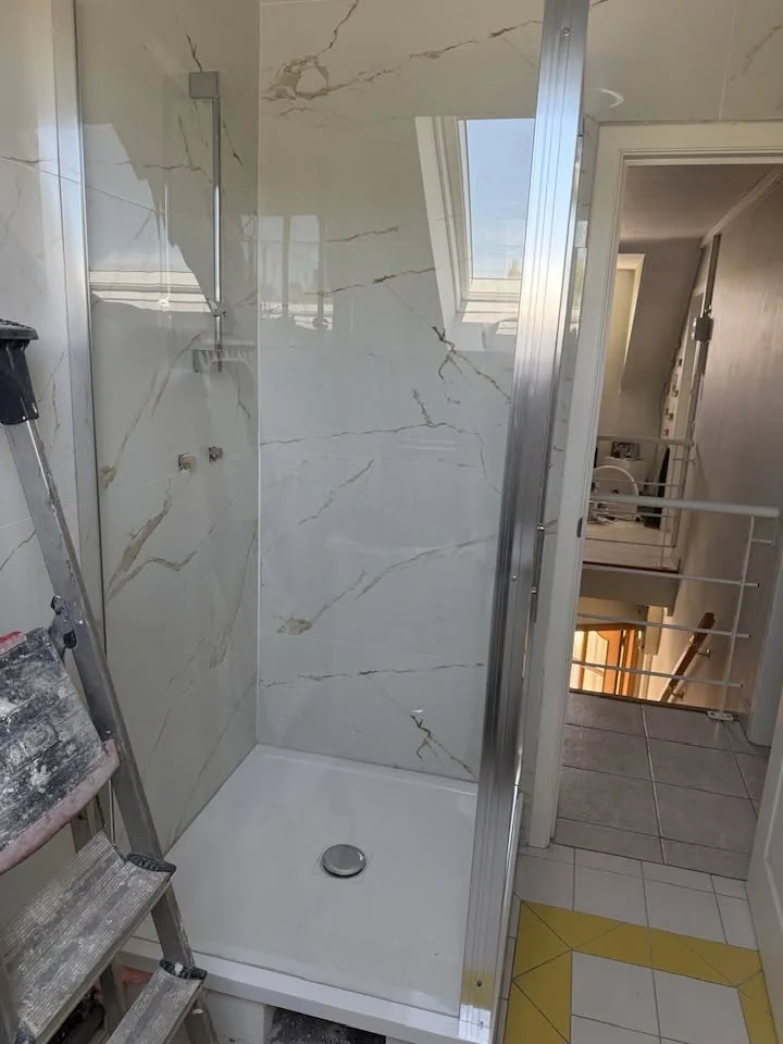Douche vitrée avec carrelage mural effet marbre, matériel de chantier visible au premier plan.