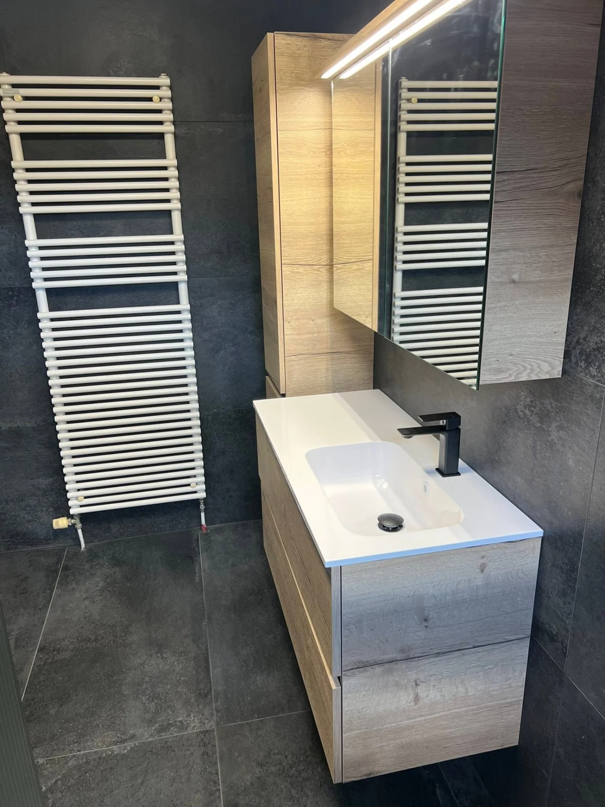 Meuble vasque avec lavabo blanc, miroir et sèche-serviettes sur un carrelage foncé.