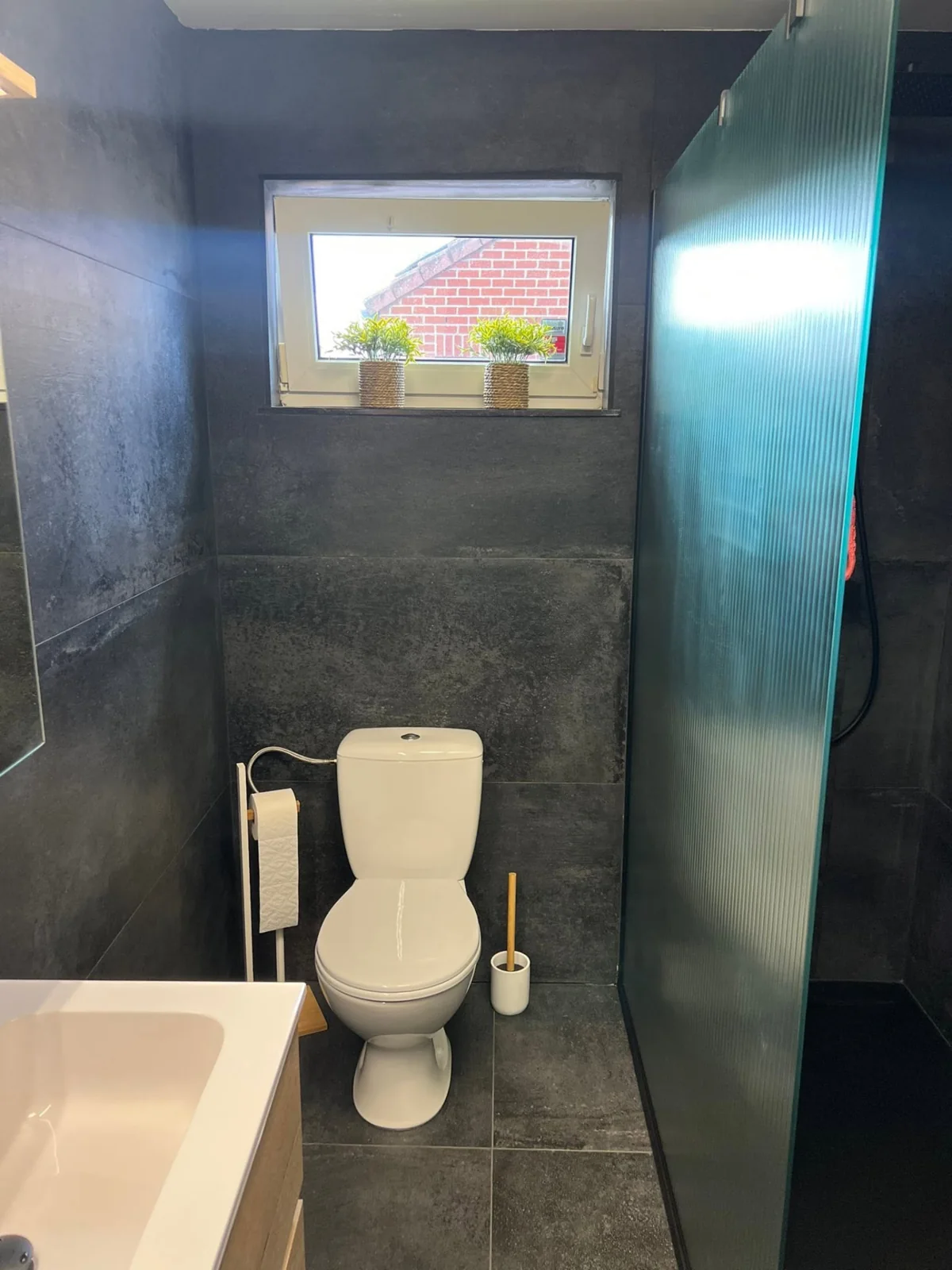 Espace WC aux murs carrelés foncés, avec fenêtre et bord du meuble vasque.